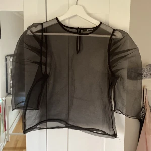 Svart transparent blus från Gina Tricot - Snygg svart transparent blus från Gina Tricot med puffiga ärmar och rund halsringning. Perfekt för en stilren och trendig look. Blusen har en knapp i nacken för enkel på- och avtagning.