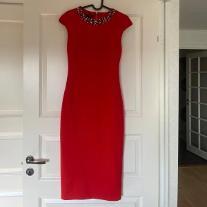 Röd klänning med pärldetaljer - Snygg röd klänning med pärldetaljer runt halsen. Den har en elegant och figurnära passform med korta ärmar. Perfekt för en festlig kväll!