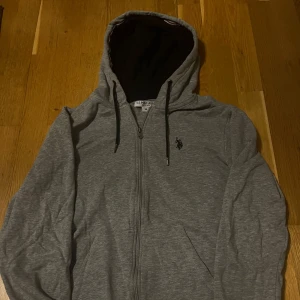 Grå hoodie från U.S. Polo Assn. - Säljer en stilren grå hoodie från U.S. Polo Assn. med dragkedja och svart logga på bröstet. Tröjan har en justerbar huva med svarta snören och är perfekt för en avslappnad look. Storlek M
