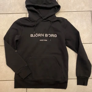 Svart hoodie från Björn Borg - Säljer en stilren svart hoodie från Björn Borg med tryck framtill. Den har en klassisk design med huva och känguruficka. Perfekt för en avslappnad look. Provad några gånger men som ny. Längd 65cm samt Bredd 45cm. Priset är PRUTBART
