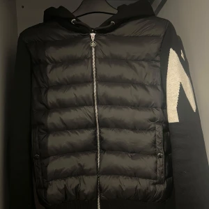 Moncler cardigan - Snygg svart jacka från Moncler med quiltad framsida och dragkedja. Jackan har en huva och långa ärmar med ett distinkt vitt mönster på sidan. Perfekt för kyligare dagar. Qr och NFC fungerar. Äkthetsbevis finns även. Vid mer frågor, skriv gärna💪🏽
