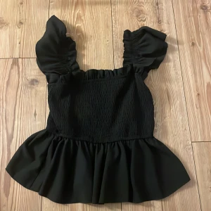 Söt Svart topp med volangdetaljer❤️ - Snygg svart topp med volangdetaljer och breda axelband. Toppen har en smockad design som ger en fin passform och en volangkant nedtill som ger extra stil. Perfekt för att piffa upp vilken outfit som helst! Knappt använd och inga defekter ❤️❤️