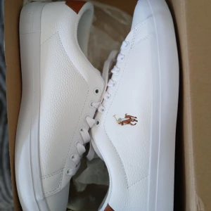 Nya Polo Ralph Lauren speakers strl 40 - Snygga vita sneakers från Polo Ralph Lauren med bruna detaljer vid hälen och klassisk logga på sidan. 100% läder. Nya