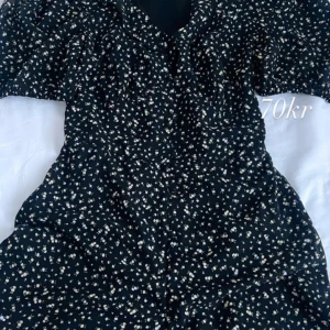 Svart blommig klänning som kopplas med short  - Säljer en svart klänning med små vita blommor. Klänningen har en v-ringad design och korta ärmar. Perfekt för en somrig look!