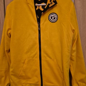 Gul och svart jacka med tryck - Säljer en cool gul och svart jacka med tryck och dragkedja. Jackan har ett unikt mönster med text och en logotyp på bröstet. Perfekt för att sticka ut i mängden!