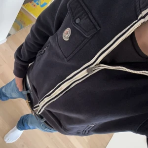Exklusiv Moncler zip hoodie - Säljer en snygg svart hoodie från Moncler med dragkedja och bröstficka. Den har långa ärmar och en klassisk logga på bröstet. Perfekt för en casual look. Köpt från merchsweden. Hör av dig vid frågor!