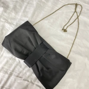 Svart kuvertväska med kedja - Elegant svart kuvertväska med en stilren kedja som axelrem. Väskan har en dekorativ rosett på framsidan som ger en chic touch. Perfekt för att lyfta din outfit till nästa nivå.