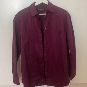 Lila skjorta från First Class Collection - Snygg lila skjorta från First Class Collection i storlek XS. Skjortan har långa ärmar och knappar framtill. Perfekt för en stilren look. Kan stajlas med t.ex en vit T-shirt under och ett par svarta kostymbyxor. Fick sy på en knapp från skjortan då den hade gått av. Hade enadast vit tråd men det är bara att byta tråd om man tycker att det är snyggare. 