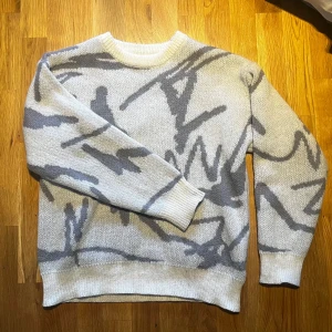 Vit och grå stickad tröja - Säljer en vit och grå stickad tröja med abstrakt mönster. Tröjan har långa ärmar och en rund halsringning. Perfekt för sval sommar kvällar!