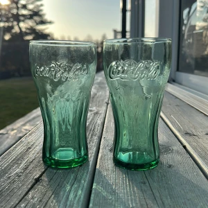 Gröna Coca-Cola glas - Säljer två klassiska gröna Coca-Cola glas med den ikoniska loggan i relief. Perfekta för att servera kalla drycker med stil. Glasen har en elegant form och är tillverkade i tjockt glas för en hållbar känsla.