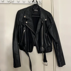 Svart skinnjacka från Zara - Snygg svart skinnjacka från Zara med dragkedjor och bälte. Jackan har en klassisk bikerstil med långa ärmar och silverfärgade detaljer.  Har storlek M. Har inte används på två år. Har väldigt bra kvalite