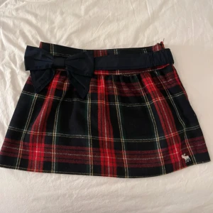 Rutig kjol från Abercrombie & Fitch - Snygg rutig kjol från Abercrombie & Fitch i rött och svart med en söt rosett framtill. Kjolen har ett klassiskt rutmönster och en liten älgbroderi i nederkanten. Perfekt för en stilren look!