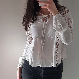 Vit blus - Säljer en söt vit blus med spetsdetaljer och knytning framtill. Blusen har långa ärmar och en luftig passform med fina hålmönster längs ärmarna och sidorna. Perfekt för en stilren look. Sitter inte tajt, kanske storlek S/M