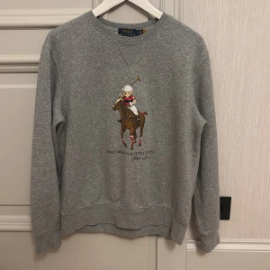 Grå sweatshirt från Polo Ralph Lauren - Säljer en grå sweatshirt från Polo Ralph Lauren med ett stort tryck av en polospelare på framsidan. Tröjan har rund hals och långa ärmar, perfekt för en avslappnad stil. Klassisk och tidlös design som funkar året runt.