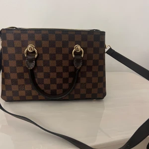 Rutig axelväska med dragkedja - Snygg rutig axelväska i brunt och svart med guldfärgade detaljer. Väskan har en praktisk dragkedja upptill och flera fack inuti för enkel organisering. Perfekt för att bära dina dagliga nödvändigheter med stil.