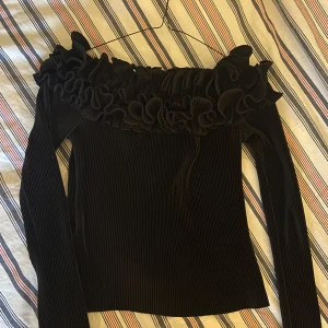Svart ribbad topp med volangdetaljer - Snygg svart ribbad topp med långa ärmar och volangdetaljer runt halsen. Perfekt för en stilren och elegant look. Köpt utomlands STORLEK XS/ S ((: