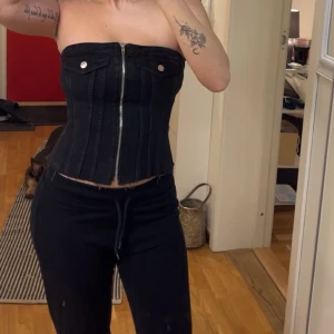 Svart jeanskorsett med dragkedja - Snygg svart korsett i jeansmaterial med dragkedja framtill och dekorativa fickor med knappar. Den är axelbandslös och har en rå kant nertill för en edgy look. Perfekt för att ge din outfit en cool touch!