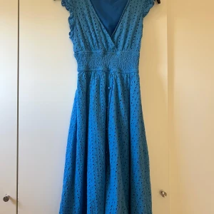 Blå klänning med broderad design - Charmig blå klänning med broderad design och omlott framtill. Den har en smickrande v-ringning och korta ärmar med volangdetaljer. Perfekt för en somrig look.