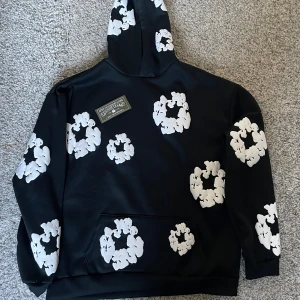 Svart hoodie med blommönster  - Säljer en svart hoodie med ett unikt vitt blommönster. Hoodien har en klassisk design med en stor ficka framtill och en bekväm huva. Perfekt för en stilren och avslappnad look. Storleken är L. Lite sämre kvalite och därav lågt pris. Hålen till fickan är är små.