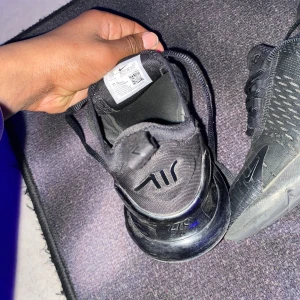 Svarta Nike Air Max 270 - Säljer ett par svarta Nike Air Max 270 sneakers med snörning. Skorna har en synlig Air-enhet i hälen och en modern design med Nike-logga på sidan. Perfekta för en sportig look.