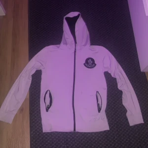 Vit hoodie från Moncler - Säljer en stilren vit hoodie från Moncler med svart dragkedja och huva. Jackan har Moncler-logga på bröstet och text på ryggen. Perfekt för en casual look. Passar S/M