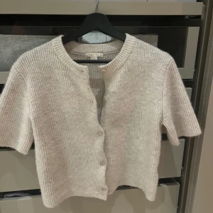 Beige stickad kofta från Gina Tricot - Säljer en mysig beige stickad kofta från Gina Tricot. Koftan har korta ärmar och knappar framtill, perfekt för lager-på-lager-stil. Den är ribbad och har en rund halsringning. Passar både till jeans och kjol för en avslappnad look.