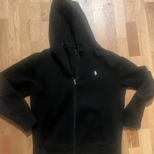 Svart hoodie från Polo Ralph Lauren - Säljer en stilren svart hoodie från Polo Ralph Lauren med dragkedja och det klassiska logotypbroderiet på bröstet. Tröjan har långa ärmar och en praktisk huva. Perfekt för en avslappnad look. Pris går såklart att diskuteras