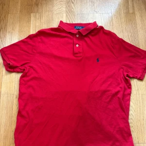 Röd Polo Ralph Lauren pikétröja XL - Klassisk röd pikétröja från Polo Ralph Lauren i bomull. Tröjan har korta ärmar, krage med två knappar och den ikoniska blå broderade loggan på bröstet. Perfekt för en clean och sportig stil.