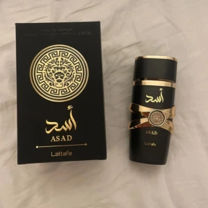 Asad parfym från Lattafa - Snygg parfymflaska från Lattafa, modell Asad. Flaskan är svart med lyxiga gulddetaljer och arabiska inskriptioner. Kommer i en matchande svart och guldig box med lejonmotiv. Perfekt för dig som gillar exklusiv design och orientaliska vibbar.