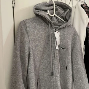 Grå hoodie från Lacoste med dragkedja - Snygg grå hoodie från Lacoste med klassisk krokodillogga på bröstet. Tröjan har huva med snörning, dragkedja framtill och en stor magficka. Tillverkad i mjuk bomullsblandning, perfekt för chill dagar. Regular fit och stilren look.