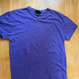 Lila t-shirt från Polo Ralph Lauren - Lila t-shirt från Polo Ralph Lauren med klassisk grön broderad logga på bröstet. T-shirten är i mjuk bomull och har en avslappnad passform med korta ärmar och rund hals. Perfekt för en chill och stilren look.