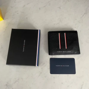 Svart plånbok Tommy Hilfiger läder - Snygg svart plånbok från Tommy Hilfiger i äkta skinn. Plånboken har två stilrena ränder i rött och vitt på framsidan samt flera kortfack och sedelfack inuti. Kommer med originalask och märkeskort. Perfekt accessoar för dig som gillar klassisk och clean stil. Mejlbekräftelse från Zalando finns