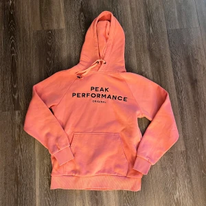 Rosa hoodie Peak Performance M - Säljer en snygg rosa hoodie från Peak Performance i storlek M. Tröjan har svart tryck framtill, huva med snörning och en stor magficka. Mjuk bomullskänsla och ribbade muddar vid ärmslut och nederkant. Perfekt för chill dagar.