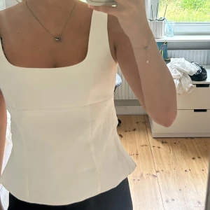 Vit linnetopp med rosett i ryggen - Superfin vit linnetopp med breda axelband, fyrkantig ringning och en stor rosett i ryggen. Toppen har en lätt utsvängd passform och öppen rygg som ger en snygg och modern look. Perfekt för varma dagar och snygg att styla med jeans eller kjol.