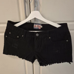 Svarta jeansshorts från Milla, strl 38 - Svarta jeansshorts från Milla med råa, fransiga kanter och klassisk femficksdesign. Shortsen har låg midja och slitna detaljer framtill för en cool, avslappnad vibe. Perfekta för sommardagar när du vill ha en edgy look.