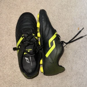 Svarta och gula fotbollsskor Pro Touch - Snygga fotbollsskor från Pro Touch i svart med ljusgröna detaljer och sula. Skorna har snörning och är tillverkade i syntetmaterial. Perfekta för dig som vill sticka ut på planen med en sportig look och bra grepp.