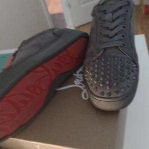 Christian Louboutin gråa  sneakers med nitar - Snygga gråa sneakers från Christian Louboutin med ikonisk röd sula och coola nitar framtill. Skorna har snörning och är tillverkade i mocka med läderdetaljer. Perfekta för dig som vill sticka ut med exklusiv streetstyle.