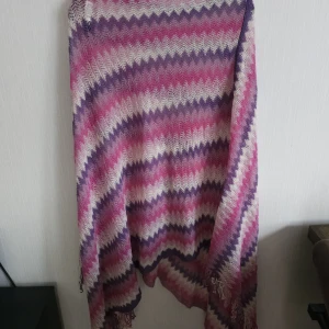 Poncho  - Mysig poncho från Miss Milla med randigt mönster i rosa, lila och vitt. Ponchon är stickad i 100% akryl och har fransar i kanterna. Perfekt att svepa runt sig för en färgglad och cool look.