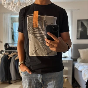 Svart t-shirt med print från Replay - Svart t-shirt från Replay med coolt tryck framtill som visar palmer och strand i gråskala, plus en orange detalj. Klassisk passform och rund hals. Perfekt för dig som gillar streetstyle och grafiska prints.