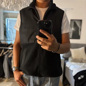 Svart fleeceväst från SOC XL - Svart väst från SOC i storlek XL med hög krage och hel dragkedja. Övre delen är i slät syntet och nedre delen i mjuk fleece. Två fickor framtill och enkel, stilren design. Perfekt lager-på-lager-plagg för kyliga dagar.