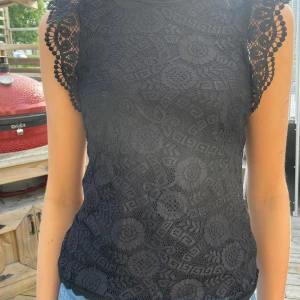 Svart spetsblus från Pieces XS - Supersnygg svart blus från Pieces i storlek XS. Blusen är täckt av detaljerad spets med blommönster och har fina virkade kanter vid ärmarna. Den är ärmlös och har en rund halsringning med knapp i nacken. Perfekt för dig som gillar en feminin och trendig look.