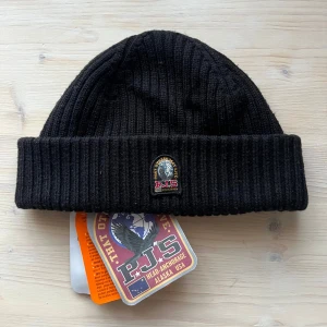 Svart ribbstickad mössa från Parajumpers - Svart ribbstickad mössa från Parajumpers med uppvikt kant och klassisk PJS-logga framtill. Tillverkad i mjukt ullmaterial som håller dig varm under kyliga dagar. Perfekt accessoar för en stilren och enkel look.
