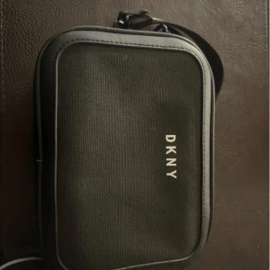 Svart axelväska från DKNY - Snygg svart axelväska från DKNY med stilren design. Väskan har ett vitt DKNY-tryck på framsidan, justerbar axelrem i svart med vit rand och cool kedjedetalj. Materialet är en mix av textil och skinnimitation. Perfekt för att bära det viktigaste med stil.