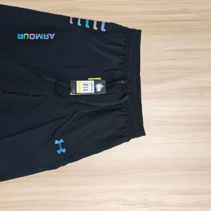 Svarta träningsshorts Under Armour - Svarta träningsshorts från Under Armour med färgglada detaljer och logga i blått. Modellen har elastisk midja med snörning och loose fit. Perfekta för sport eller gym och tillverkade i lätt syntetmaterial som andas.