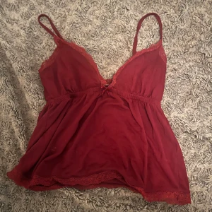 Vinröd linnetopp med spetsdetaljer - Söt vinröd linnetopp med tunna axelband och spets längs urringning och nederkant. Toppen har en liten rosett framtill och är lätt utsvängd för en avslappnad look.