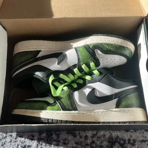 Nike Air Jordan 1 Low svart/grön 38,5 - Snygga Nike Air Jordan 1 Low med svart, vitt och grönt läder. Skorna har en unik grön textur på panelerna, svarta detaljer och limegröna skosnören. Klassisk Jordan-logga på hälen och svart swoosh på sidorna. Perfekta för dig som vill sticka ut.
