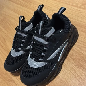 Svarta Dior b22  - Chunky svarta sneakers från Dior med silvriga reflexdetaljer och meshpaneler. Skorna har snörning, grov sula och coola kontraster mellan material. Perfekta för dig som vill sticka ut med en sportig och modern vibe.