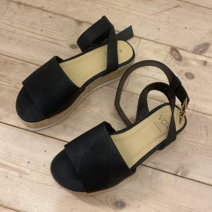 Svarta platåsandaler från VOX - Snygga svarta sandaler från VOX med breda remmar och guldfärgat spänne. Skorna har en hög platåsula i flätad jute-look och vit räfflad undersida. Veganskt material och öppen tå gör dem perfekta för varma dagar.