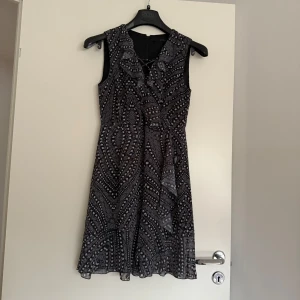 Svart mönstrad kortklänning Guess - Supersnygg svart kortklänning från Guess med vitt och grått grafiskt mönster. Ärmlös modell med lätt v-ringning och volangdetalj framtill. Klänningen har en feminin passform och är tillverkad i ett tunt, flowy material som känns lyxigt.