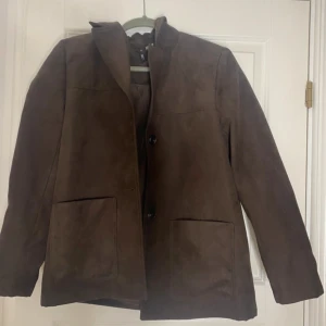 Brun mocka kavaj från H&M, stl S - Snygg brun kavaj i mockaimitation från H&M. Jackan har två stora fickor framtill, knäppning med två knappar och klassisk krage. Perfekt för dig som gillar stilrena och tidlösa plagg med en modern touch.